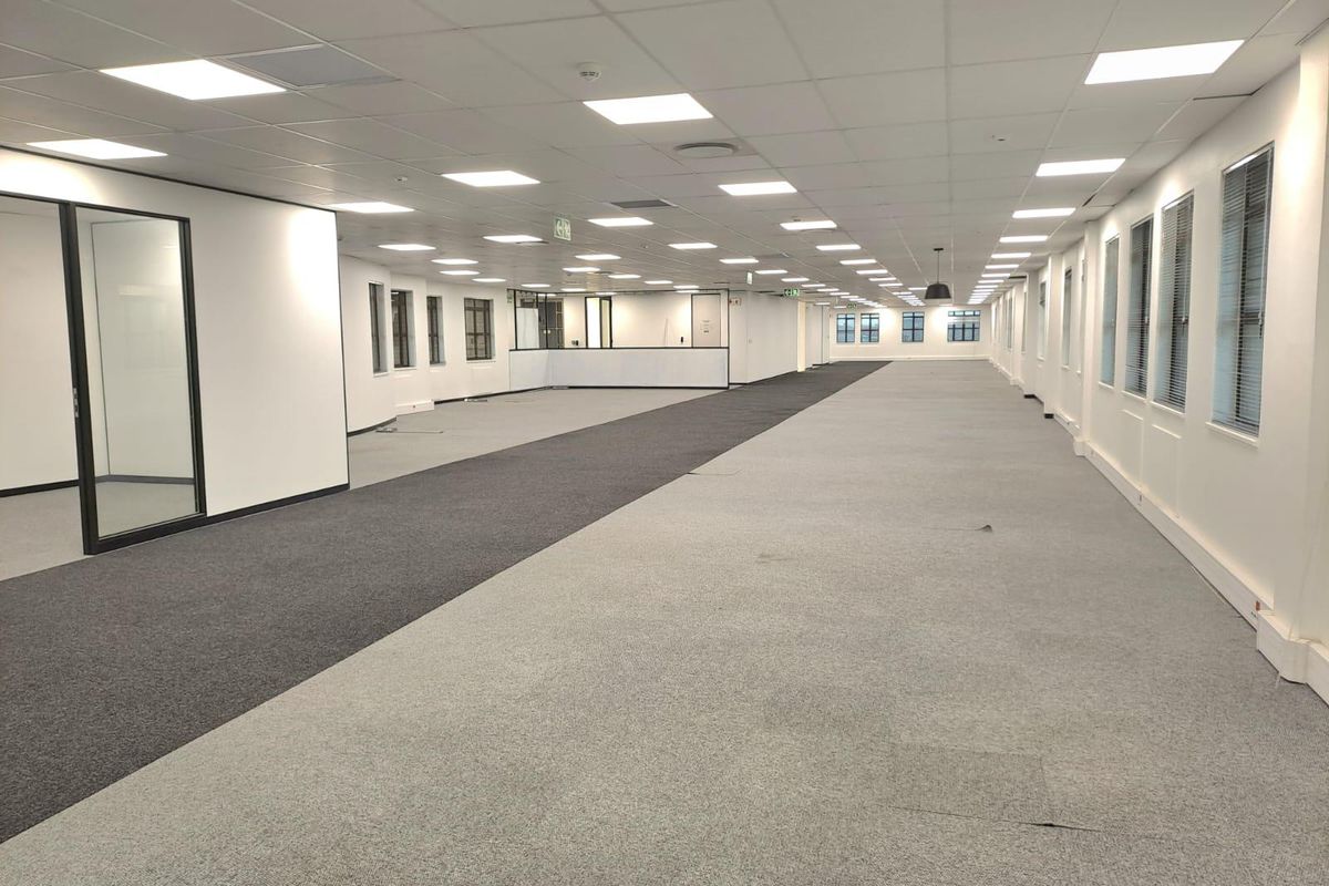 Spacious open-plan office area featuring recessed ceiling lighting, large windows with blinds, and glass-partitioned office space.