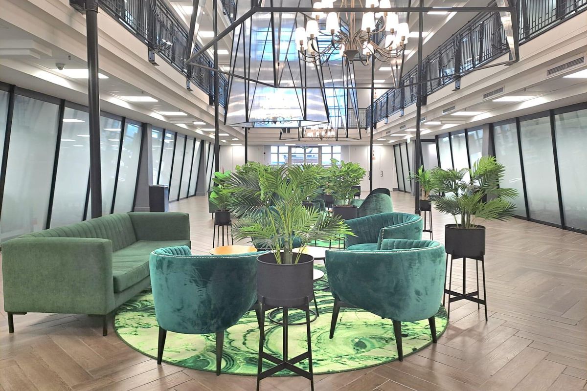 A spacious, modern open-plan office atrium featuring high ceilings, herringbone wood-look flooring, and a mezzanine level with black metal railings.