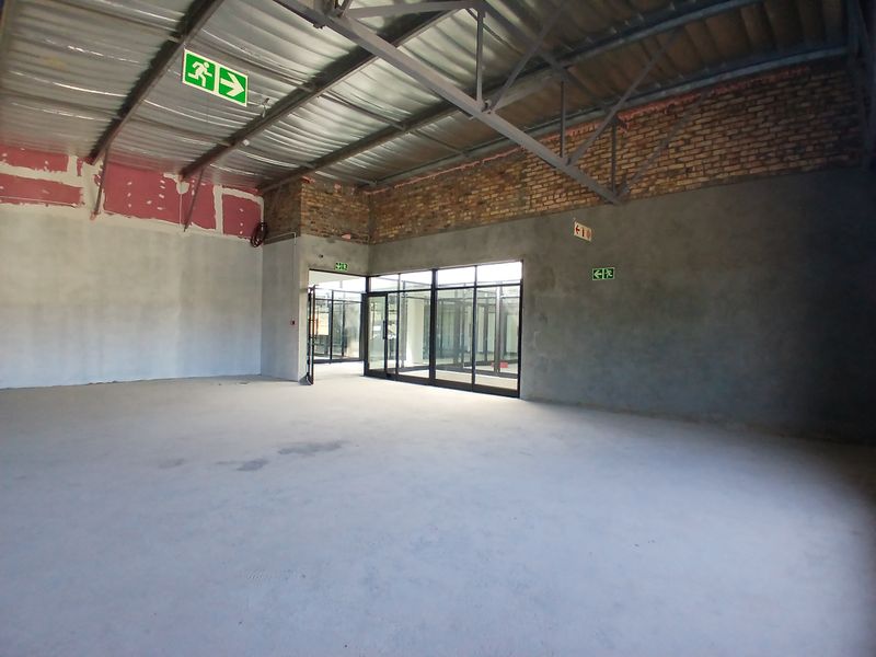 An expansive, unfinished open-plan commercial space featuring exposed steel roof trusses, raw brickwork, cement flooring, and modern aluminium-framed glass partitioning.