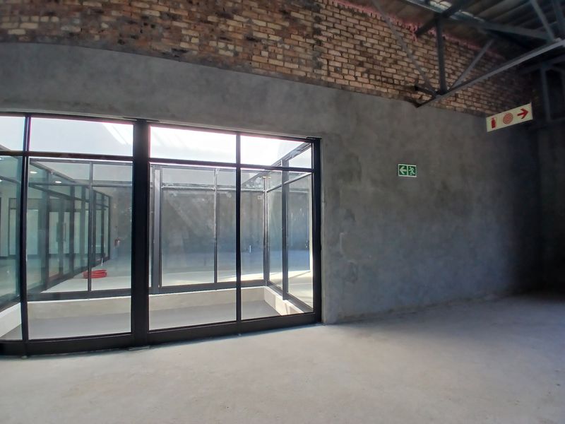 An expansive, unfinished open-plan commercial space featuring exposed brick walls, steel roof trusses, concrete flooring, and large glass partitions.