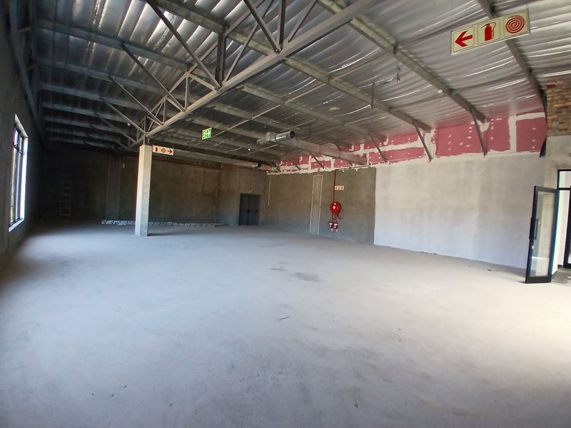 A large, open-plan commercial space featuring exposed steel roof trusses, concrete flooring, and industrial-style ventilation, currently in an unfinished shell condition.