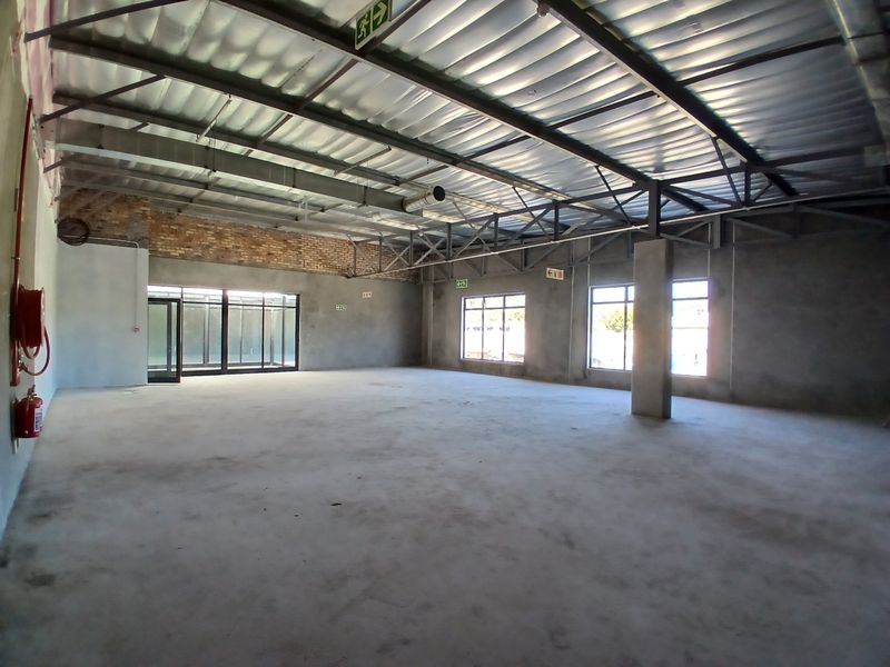 A spacious, open-plan commercial interior featuring exposed steel roof trusses, concrete flooring, and large windows, currently in an unfinished shell condition.