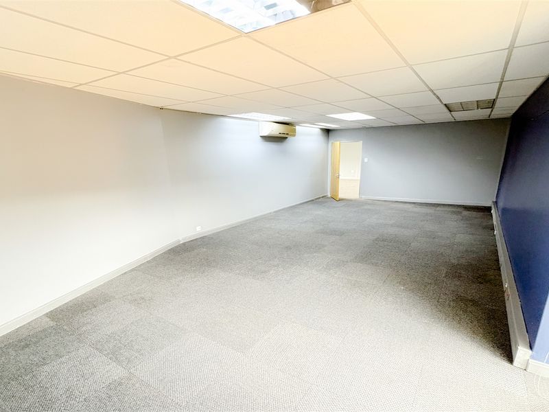 A spacious, empty office area featuring carpeted flooring, a suspended ceiling with recessed lighting, and a wall-mounted air conditioning unit.