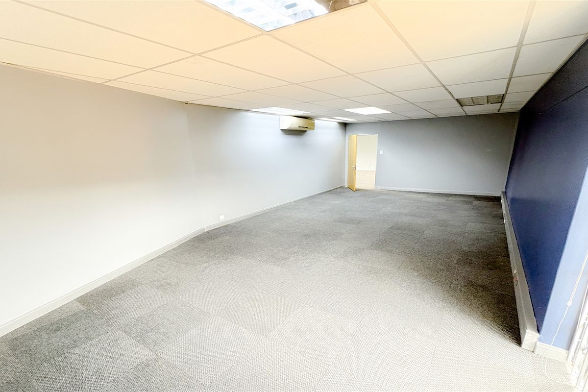 A spacious, empty office area featuring carpeted flooring, a suspended ceiling with recessed lighting, and a wall-mounted air conditioning unit.