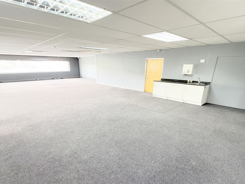 Spacious open-plan office area featuring a suspended ceiling, recessed lighting, carpeted flooring, and a convenient built-in kitchenette with a sink.