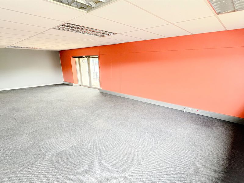 A spacious, empty office area featuring carpet tiles, a suspended ceiling with recessed lighting, and a glass sliding door providing natural light.
