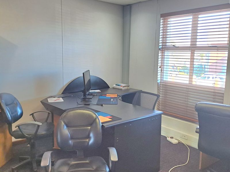 A functional office space featuring carpeted flooring, a suspended ceiling with recessed lighting, and a window fitted with venetian blinds.