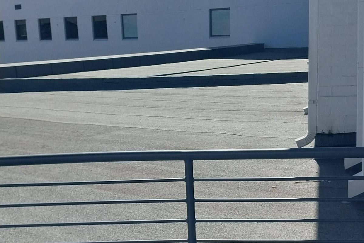 An elevated view of a flat roof section with a metal safety railing, adjacent to a white painted office building facade.