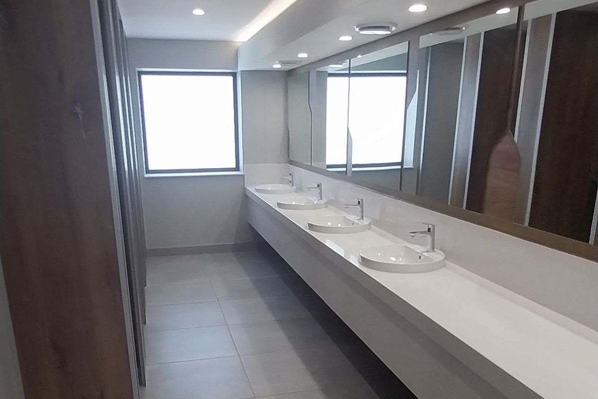 Modern office restroom featuring a long vanity with multiple integrated sinks, large wall-to-wall mirrors, recessed ceiling lighting, and tiled flooring.