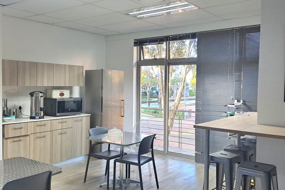 A functional office kitchenette area featuring light wood cabinetry, laminate flooring, and a large window with blinds providing natural light and exterior views.