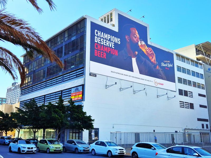 Exterior view of a multi-story office building featuring a prominent billboard, street-level parking, and security fencing in an urban setting.