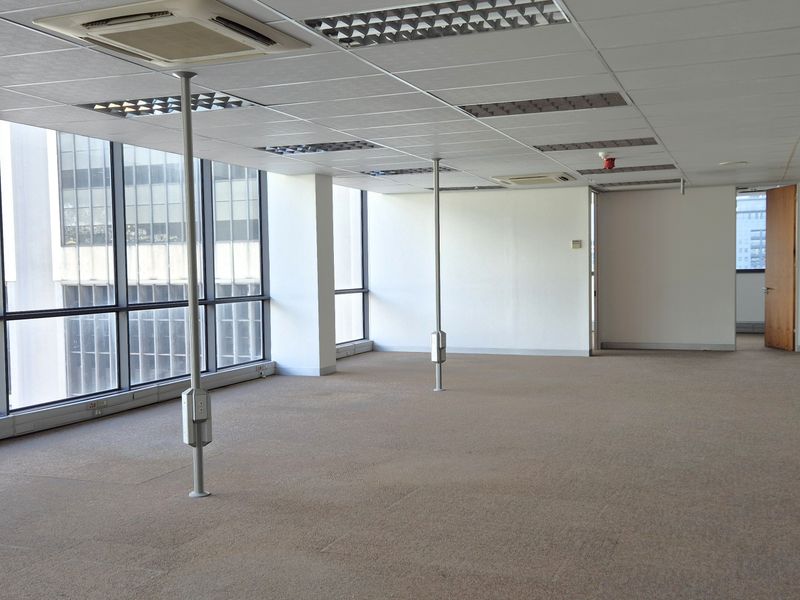 A spacious, empty open-plan office area featuring large floor-to-ceiling windows, suspended ceilings with recessed lighting, and carpeted flooring.