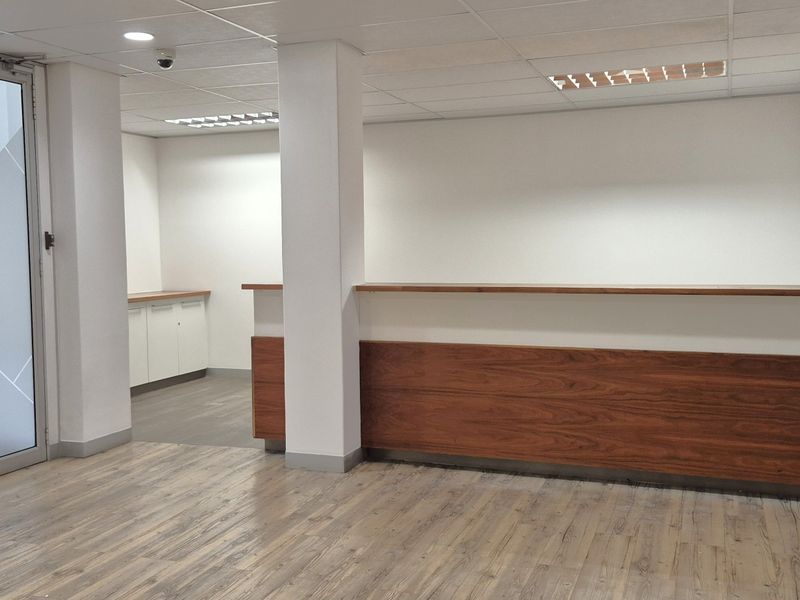 A clean, modern office reception area featuring a built-in wooden reception desk, laminate flooring, and a glass entrance door.