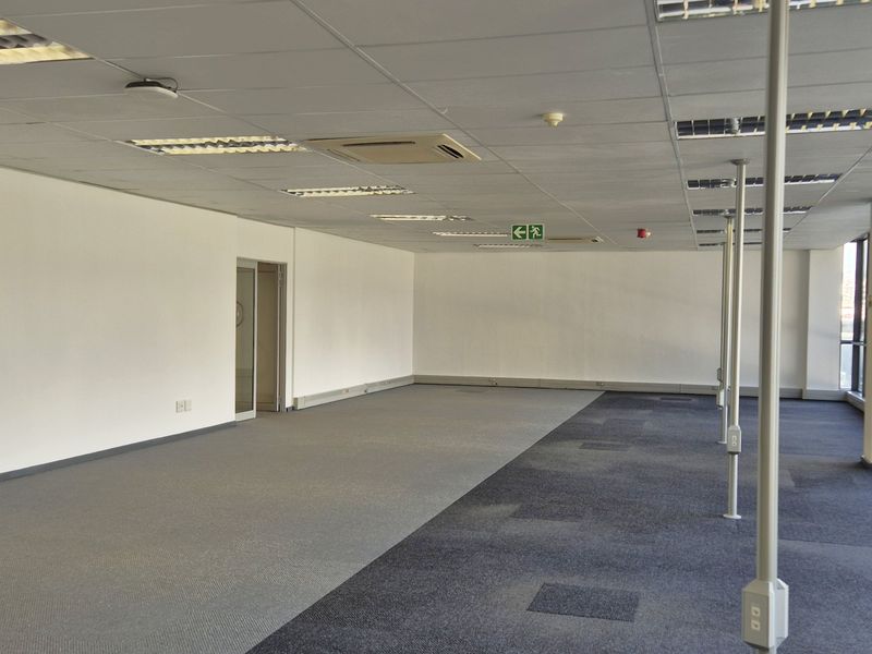 A spacious, open-plan office area featuring large floor-to-ceiling windows, carpeted flooring, and a suspended ceiling with integrated lighting and air conditioning.