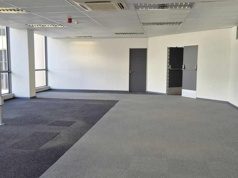 A spacious, vacant open-plan office featuring large windows, suspended ceilings with integrated lighting, and carpeted flooring.