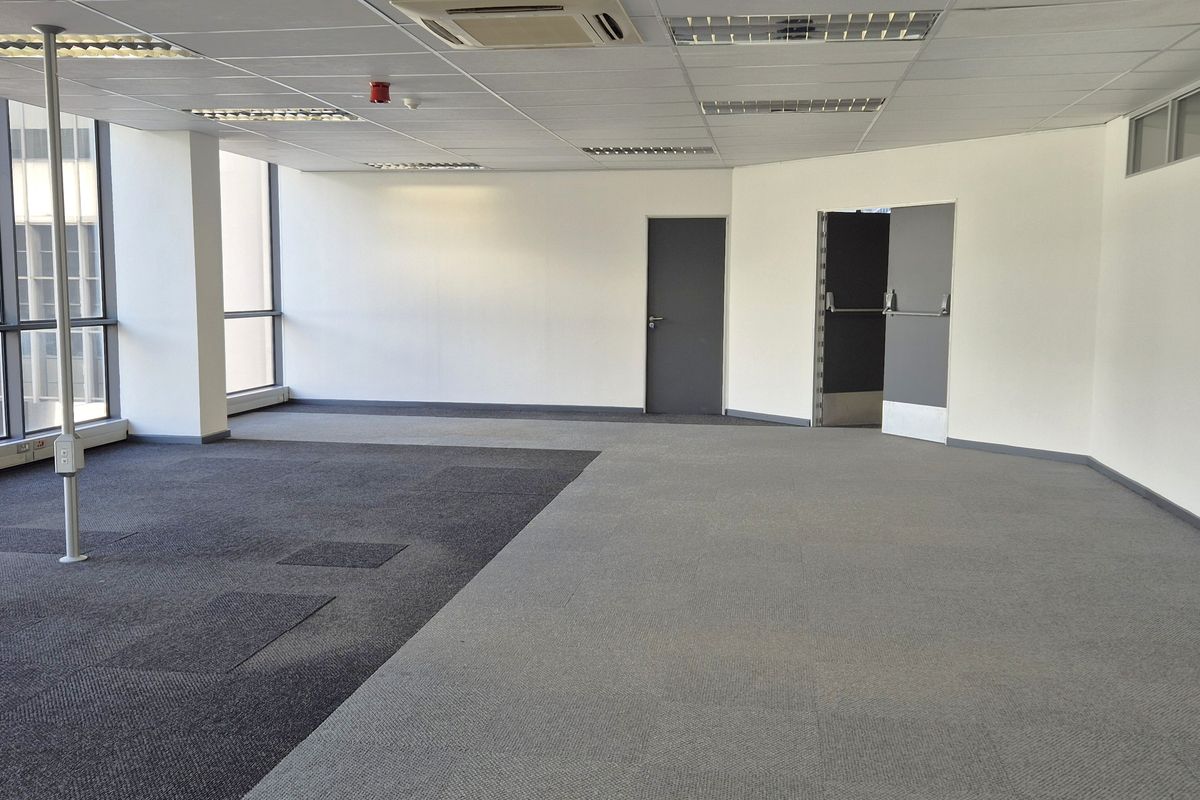 A spacious, vacant open-plan office featuring large windows, suspended ceilings with integrated lighting, and carpeted flooring.