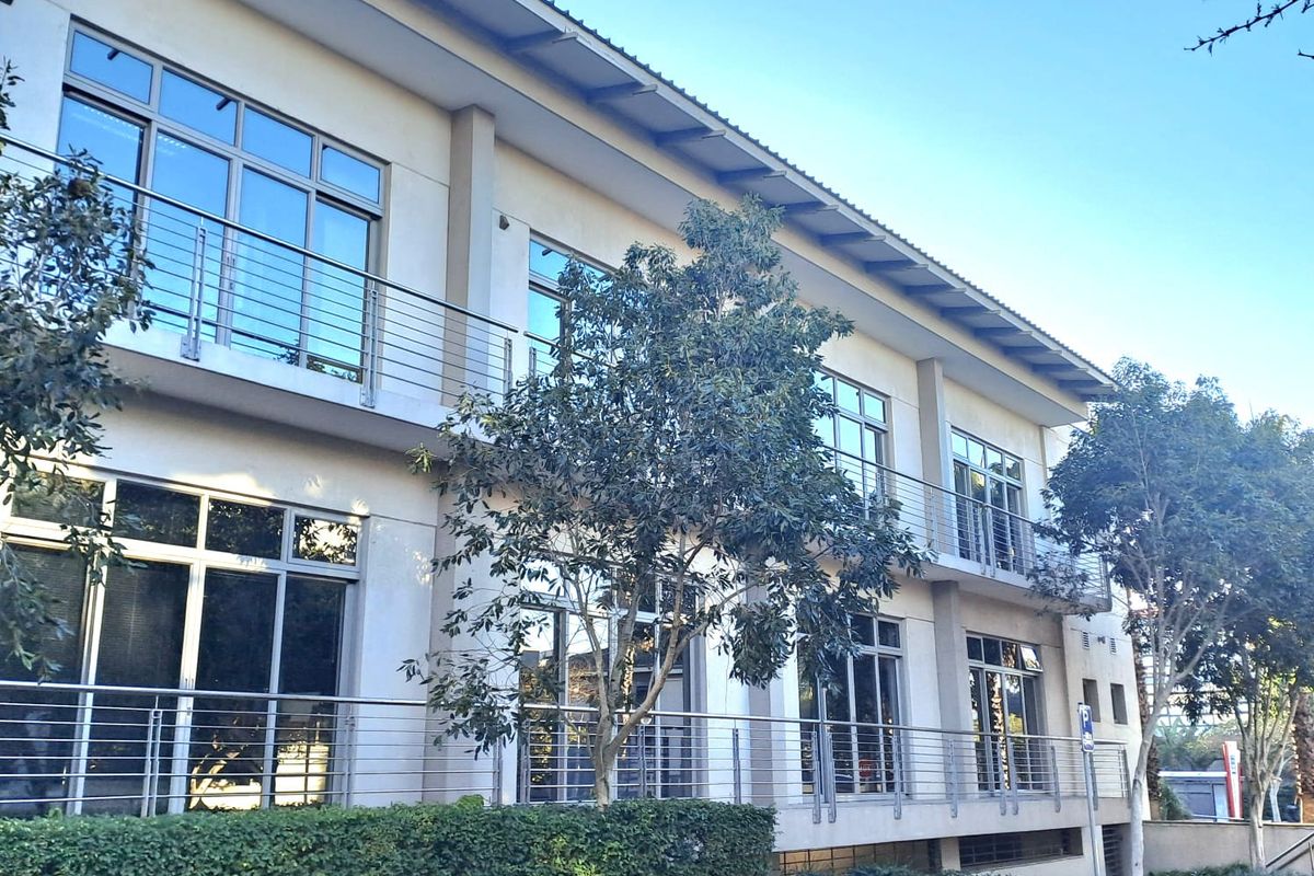 A modern two-story office building featuring large glass windows, metal balcony railings, and a paved exterior walkway with landscaped garden beds.
