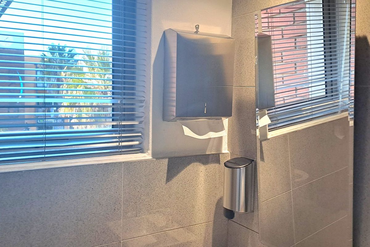 A modern office restroom featuring grey tiled walls, a wall-mounted basin with a chrome faucet, a mirror, and stainless steel paper towel and soap dispensers.