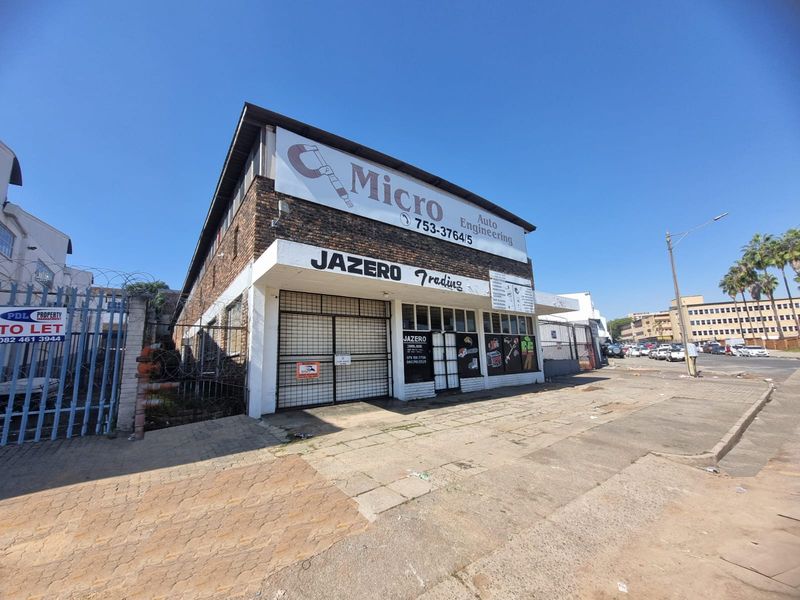 Street-level view of a commercial retail property featuring brick facade, security gates, and prominent signage, situated along a paved sidewalk.