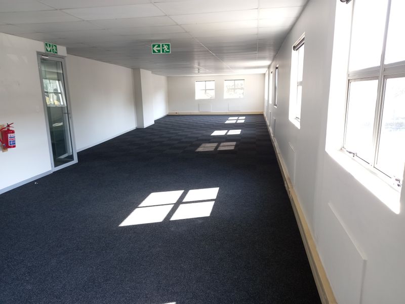 A spacious, empty open-plan office area featuring carpeted flooring, suspended ceilings, and large windows providing ample natural light.