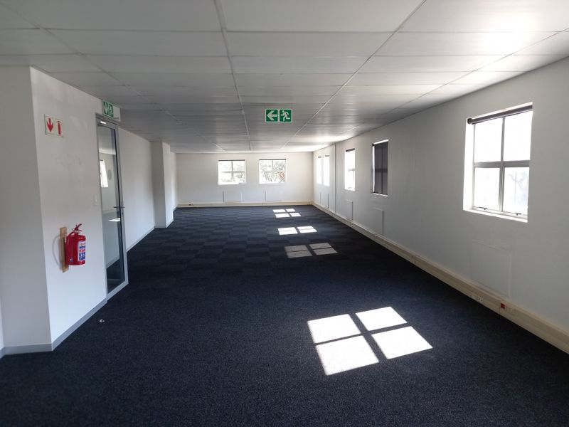 A spacious, empty open-plan office area featuring carpeted flooring, suspended ceilings, and multiple windows providing natural light.