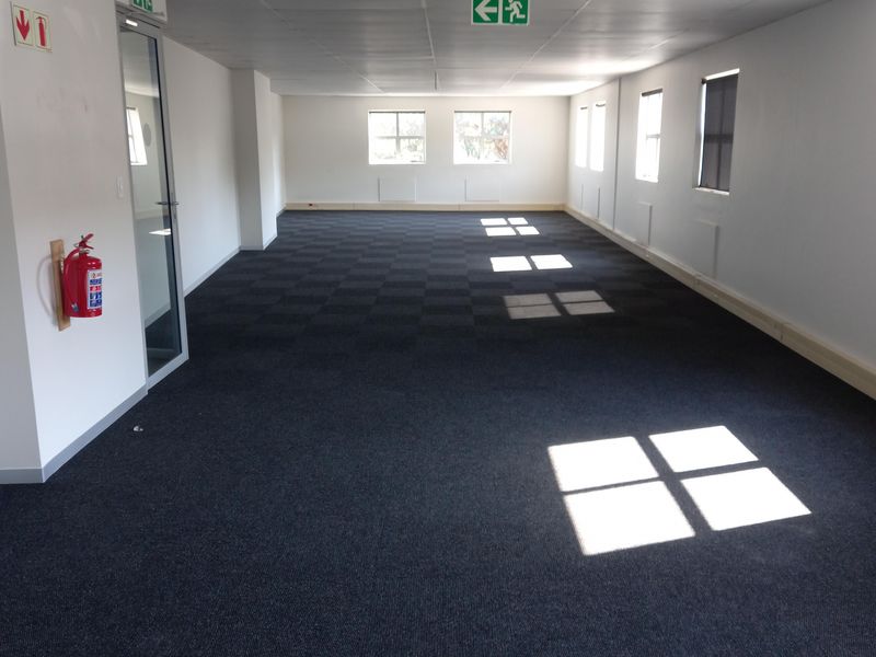 A spacious, empty open-plan office area featuring dark carpet tiles, a suspended ceiling, and natural light from multiple windows.