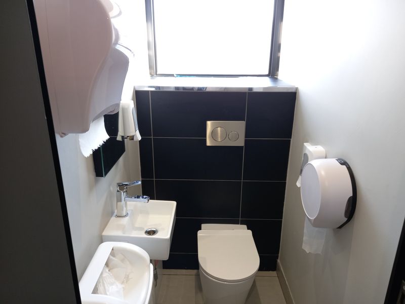 A compact modern office restroom featuring a wall-mounted toilet with a concealed cistern, a small basin with a chrome mixer tap, and dark tiling.