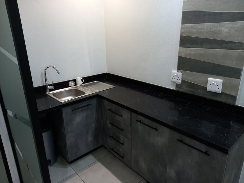 A modern, compact office kitchenette featuring sleek grey cabinetry, a black stone countertop, stainless steel sink, and contemporary tiled splashback.