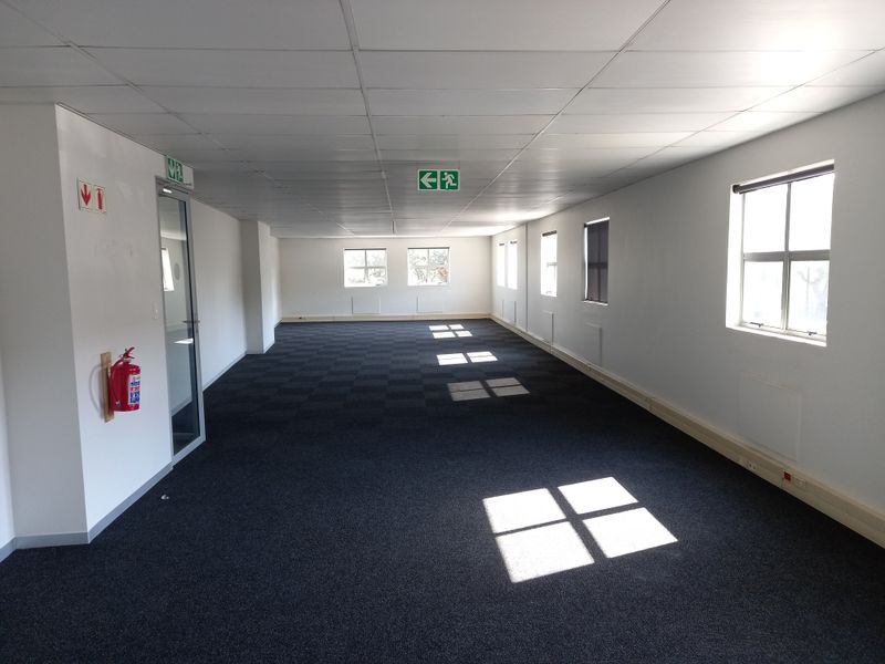 A spacious, empty open-plan office area featuring carpeted flooring, suspended ceilings, and multiple windows providing natural light.