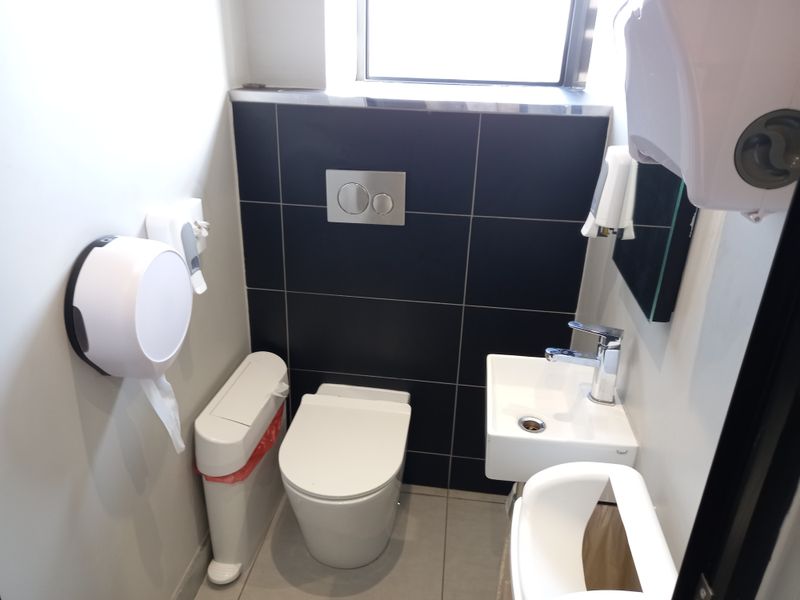 A compact modern office restroom featuring a wall-mounted toilet with concealed cistern, a small basin with chrome mixer tap, and dark tiled wall accents.