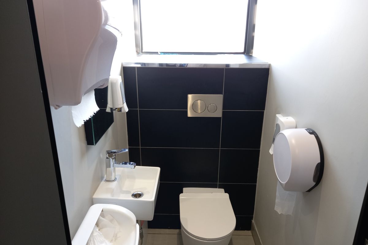 A compact modern office restroom featuring a wall-mounted toilet with a concealed cistern, a small basin with a chrome mixer tap, and dark tiling.