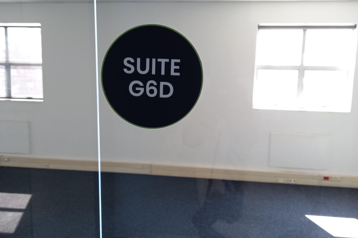 An empty office suite featuring carpeted flooring, a suspended ceiling, and a glass partition wall with suite signage.
