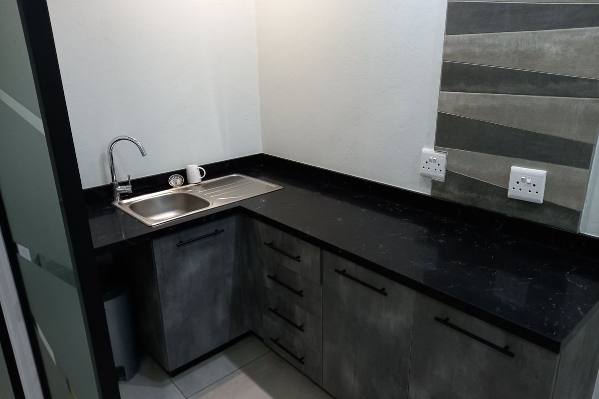 A modern, compact office kitchenette featuring sleek grey cabinetry, a black stone countertop, stainless steel sink, and contemporary tiled splashback.