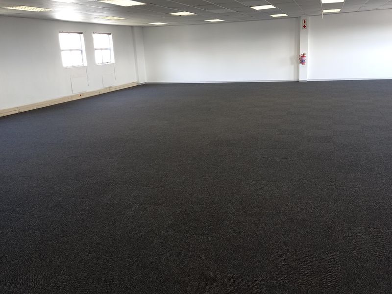 A spacious, open-plan office area featuring neutral carpet tiles, a suspended ceiling with recessed lighting, and a wall-mounted fire extinguisher.