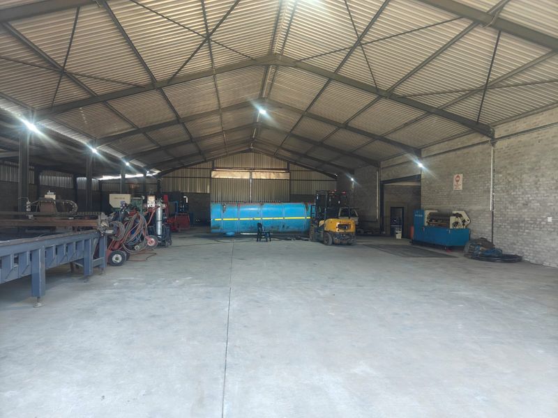 Large industrial warehouse or workshop space featuring high-clearance corrugated metal roofing, exposed steel trusses, and durable concrete flooring.