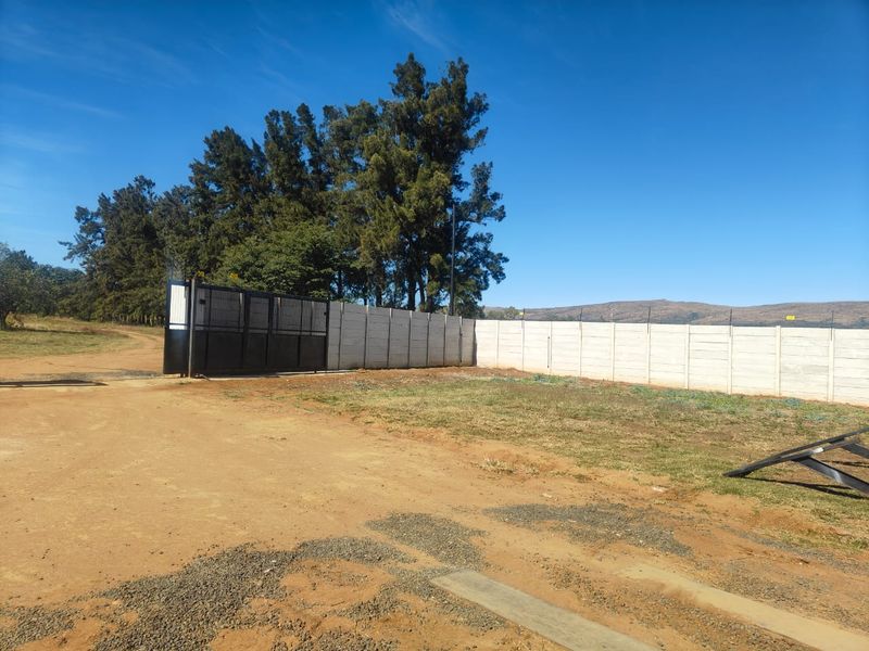 A view of the property entrance featuring a secure metal sliding gate and extensive precast concrete boundary walling on a dirt access road.