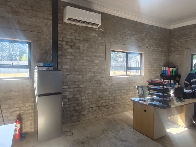 A functional office space featuring exposed brick walls, concrete flooring, and a wall-mounted air conditioning unit with natural light from aluminium-framed windows.