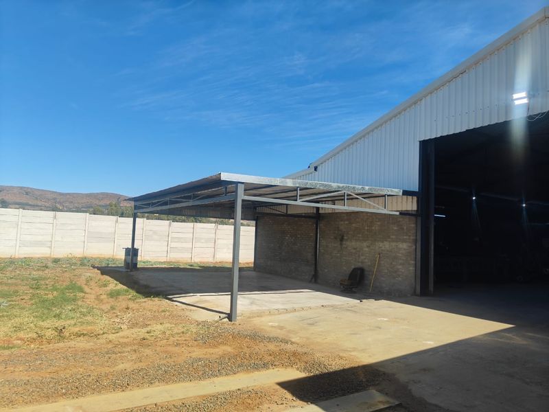 An outdoor covered workspace or storage area featuring a steel-framed roof structure, concrete flooring, and adjacent industrial building access.