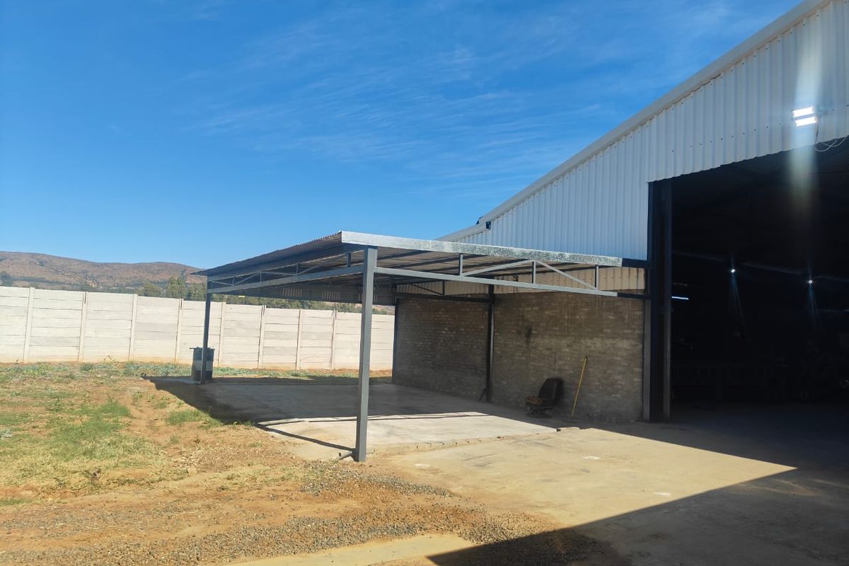 An outdoor covered workspace or storage area featuring a steel-framed roof structure, concrete flooring, and adjacent industrial building access.