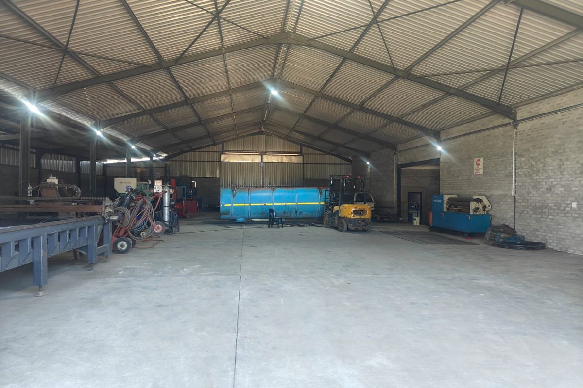 Large industrial warehouse or workshop space featuring high-clearance corrugated metal roofing, exposed steel trusses, and durable concrete flooring.