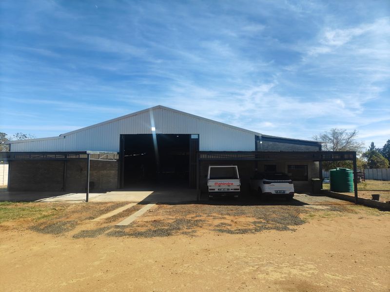Large industrial-style shed or workshop building featuring a corrugated metal roof, steel frame construction, and integrated covered parking bays on a small holding.