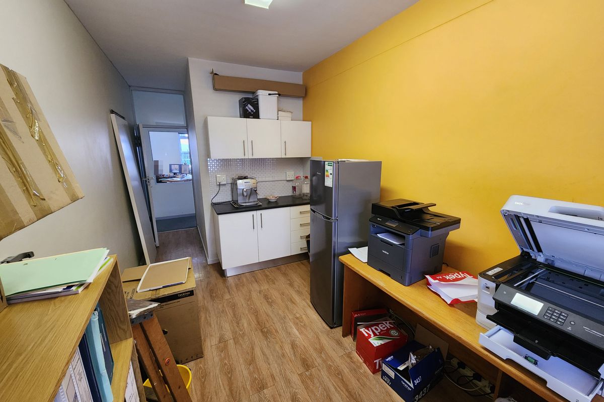 A functional office kitchenette featuring white cabinetry, tiled backsplash, and wood-look laminate flooring, currently utilized as a breakroom and storage area.