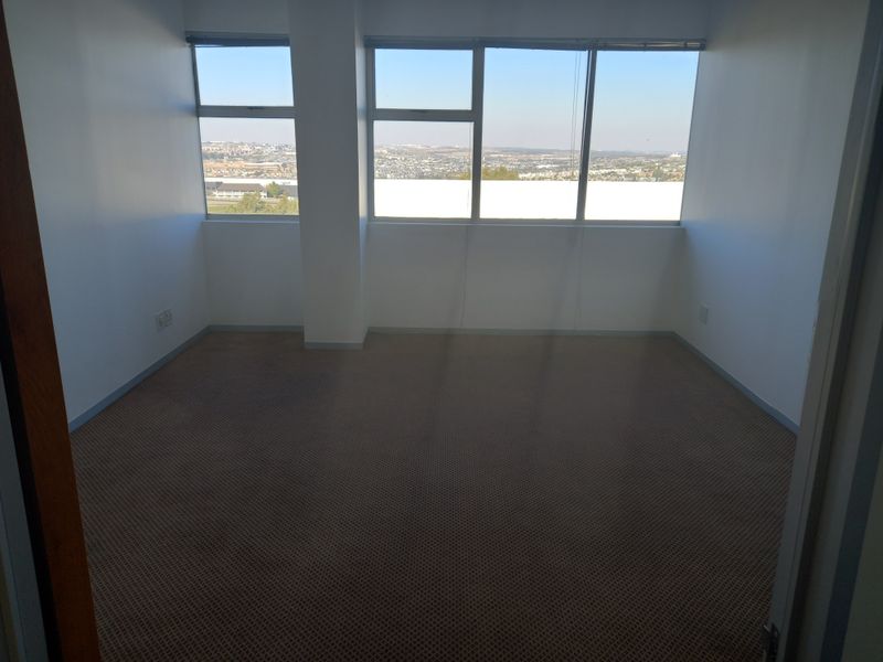 A bright, empty office space featuring large windows with expansive city views and neutral carpeted flooring.