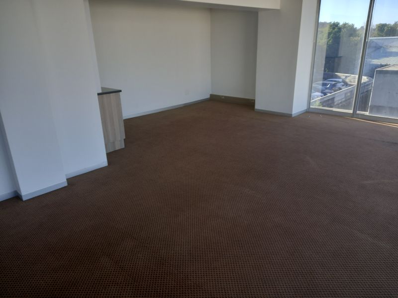 An empty office space featuring brown carpet flooring, a small built-in kitchenette counter, and large windows providing natural light.