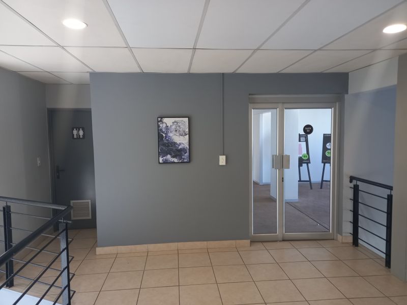 A clean office hallway featuring tiled flooring, a suspended ceiling with recessed lighting, and a glass entrance door leading to an office suite.