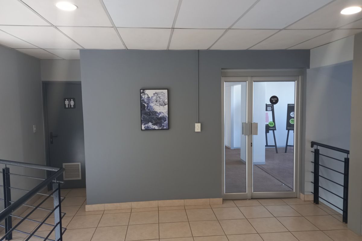 A clean office hallway featuring tiled flooring, a suspended ceiling with recessed lighting, and a glass entrance door leading to an office suite.