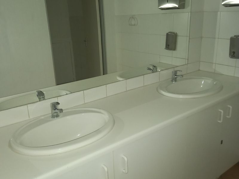 A functional office bathroom featuring a double vanity with ceramic sinks, chrome faucets, a large wall mirror, and tiled flooring.