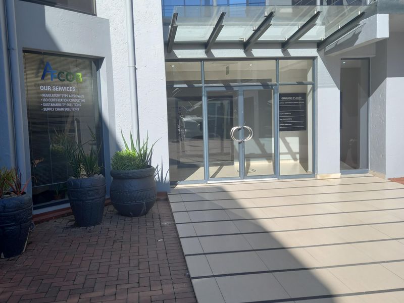 Modern office building entrance featuring a glass canopy, aluminium framed glass doors, and a combination of paved and tiled exterior flooring.