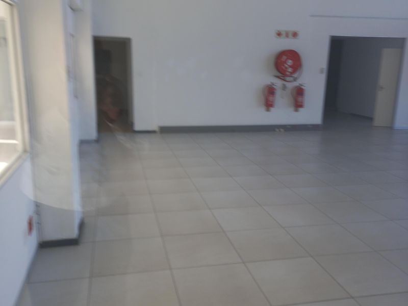 A spacious, empty office area featuring tiled flooring, a suspended ceiling, and wall-mounted fire safety equipment.