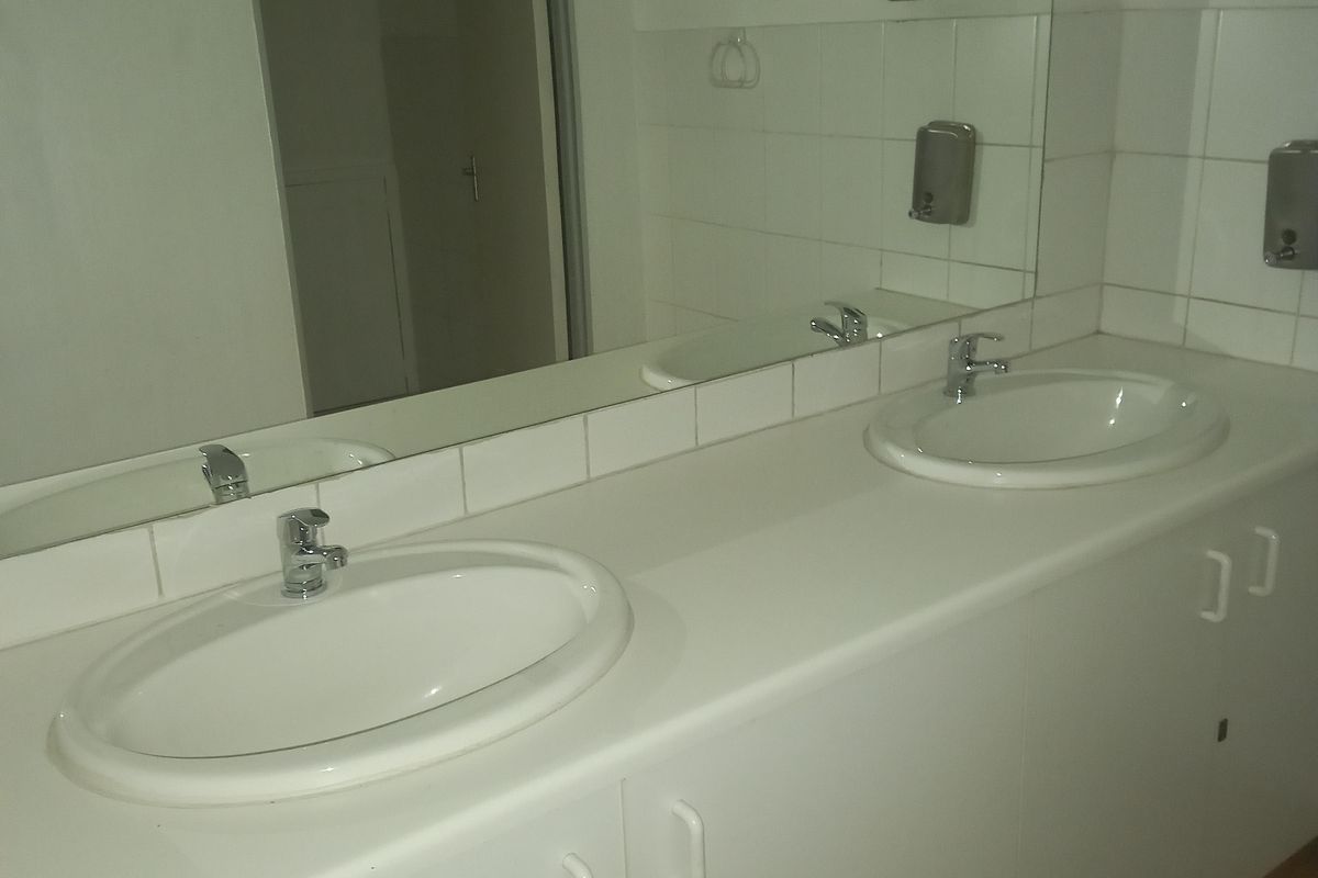 A functional office bathroom featuring a double vanity with ceramic sinks, chrome faucets, a large wall mirror, and tiled flooring.